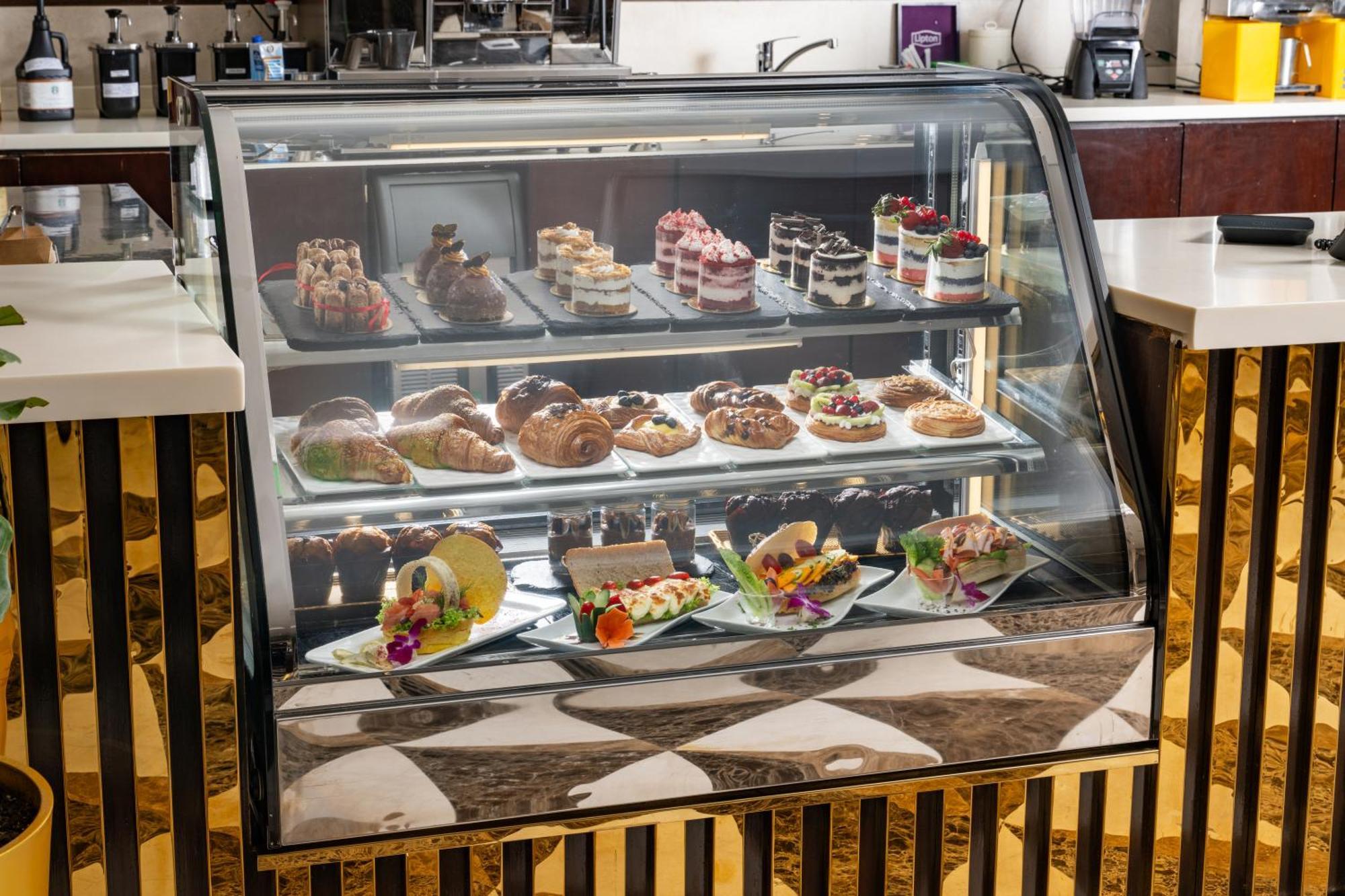 Al Aqeeq Madinah Hotel Medyna Zewnętrze zdjęcie The photo shows a display case filled with an assortment of desserts. There are various types of pastries, including cakes, tarts, and croissants, arranged neatly on different shelves. The desserts are visually appealing, with colorful decorations, f