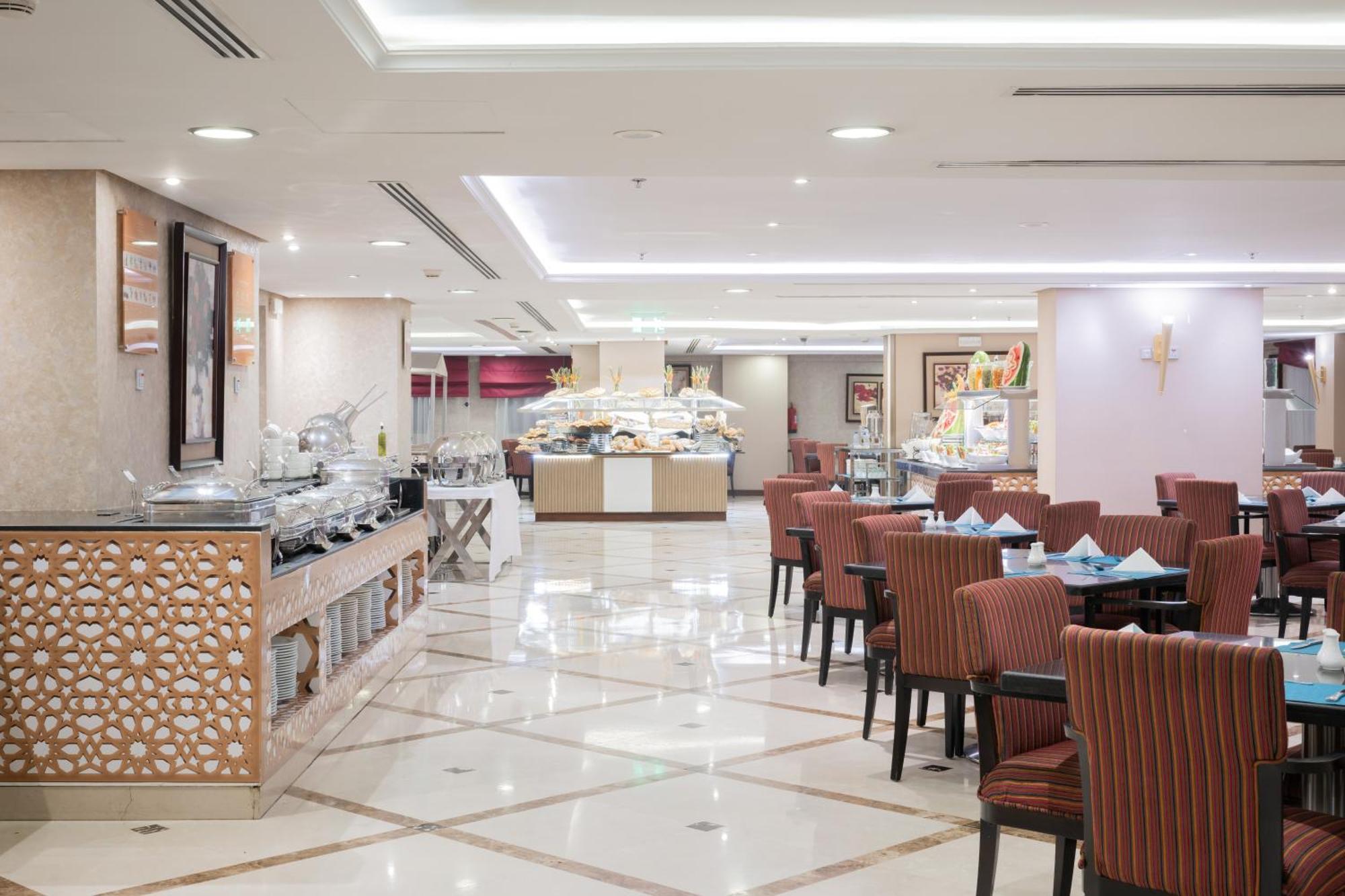 Al Aqeeq Madinah Hotel Medyna Zewnętrze zdjęcie The photo shows an elegantly designed restaurant or dining area. It features a spacious layout with a glossy tiled floor and bright, soft lighting. There are several dining tables with striped chairs arranged neatly. Along one side, there's a buffet 