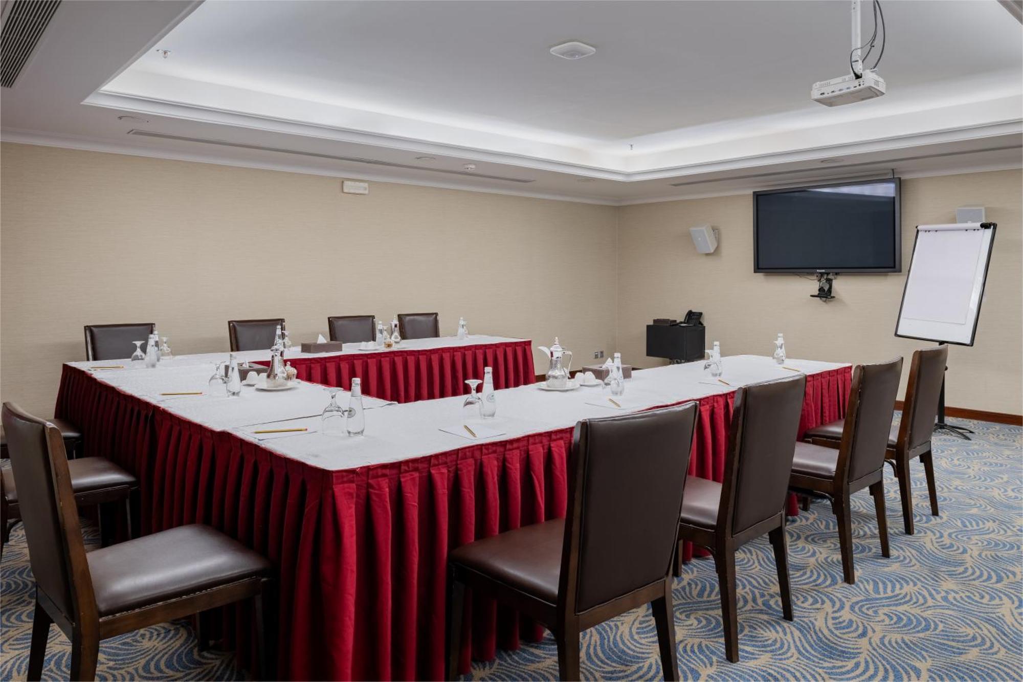 Al Aqeeq Madinah Hotel Medyna Zewnętrze zdjęcie The image shows a conference room set up for a meeting. There is a U-shaped arrangement of tables covered with white linens and red table skirts. Each table position is equipped with a water bottle and a small notepad, suggesting that it is designed 