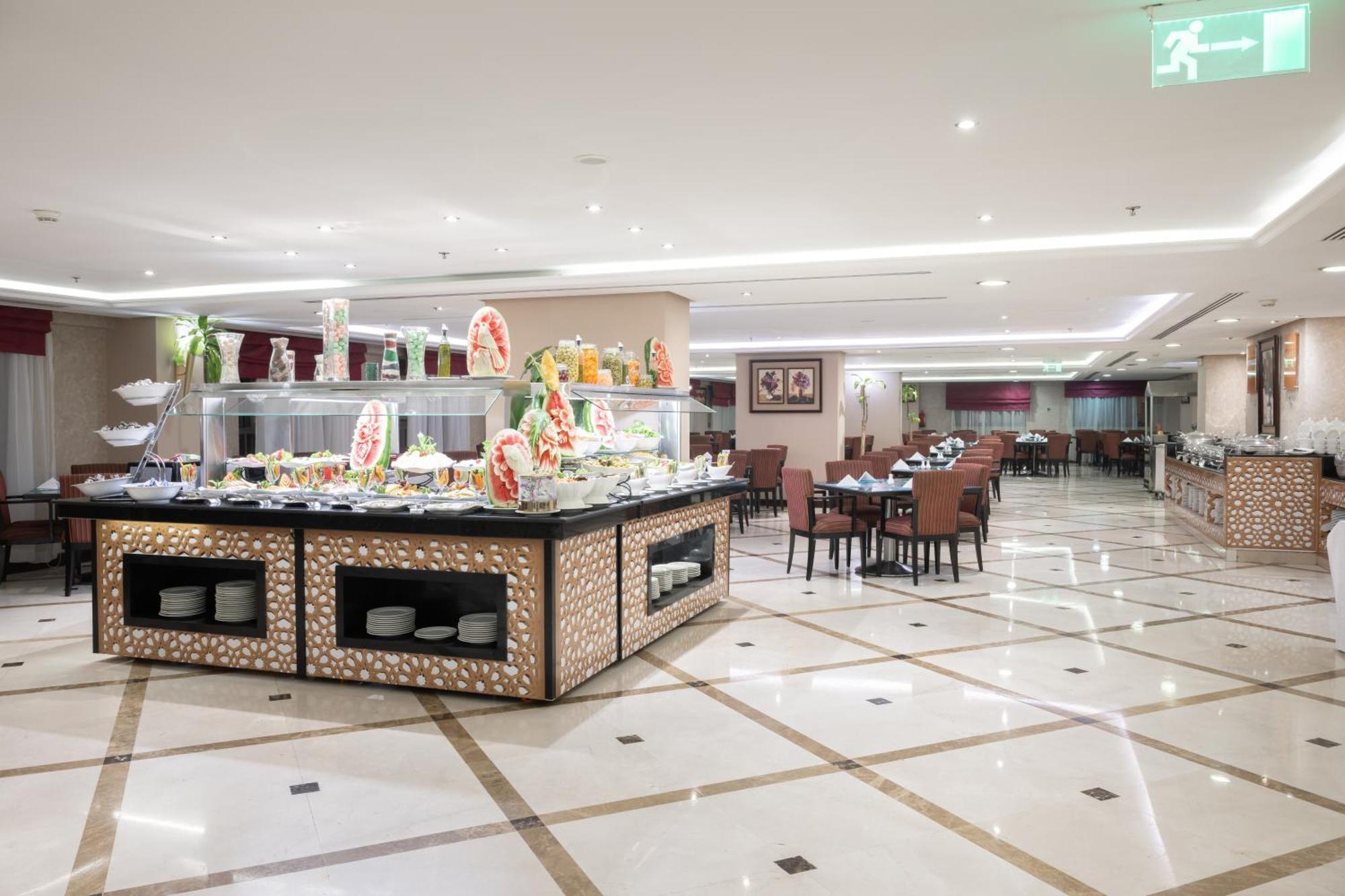 Al Aqeeq Madinah Hotel Medyna Zewnętrze zdjęcie The photo shows a spacious restaurant interior with a modern design. In the foreground, there is a large buffet counter filled with various food items, beautifully arranged and decorated. The counter features a backlit display, showcasing fruits and 