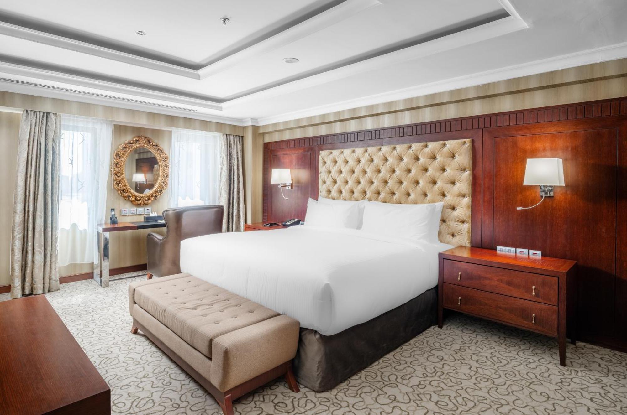 Al Aqeeq Madinah Hotel Medyna Zewnętrze zdjęcie The photo shows a well-furnished hotel room featuring a large bed with a tufted headboard. The room has a warm color palette with wooden accents and soft lighting. There are two windows with curtains that allow natural light to fill the space. Alongs
