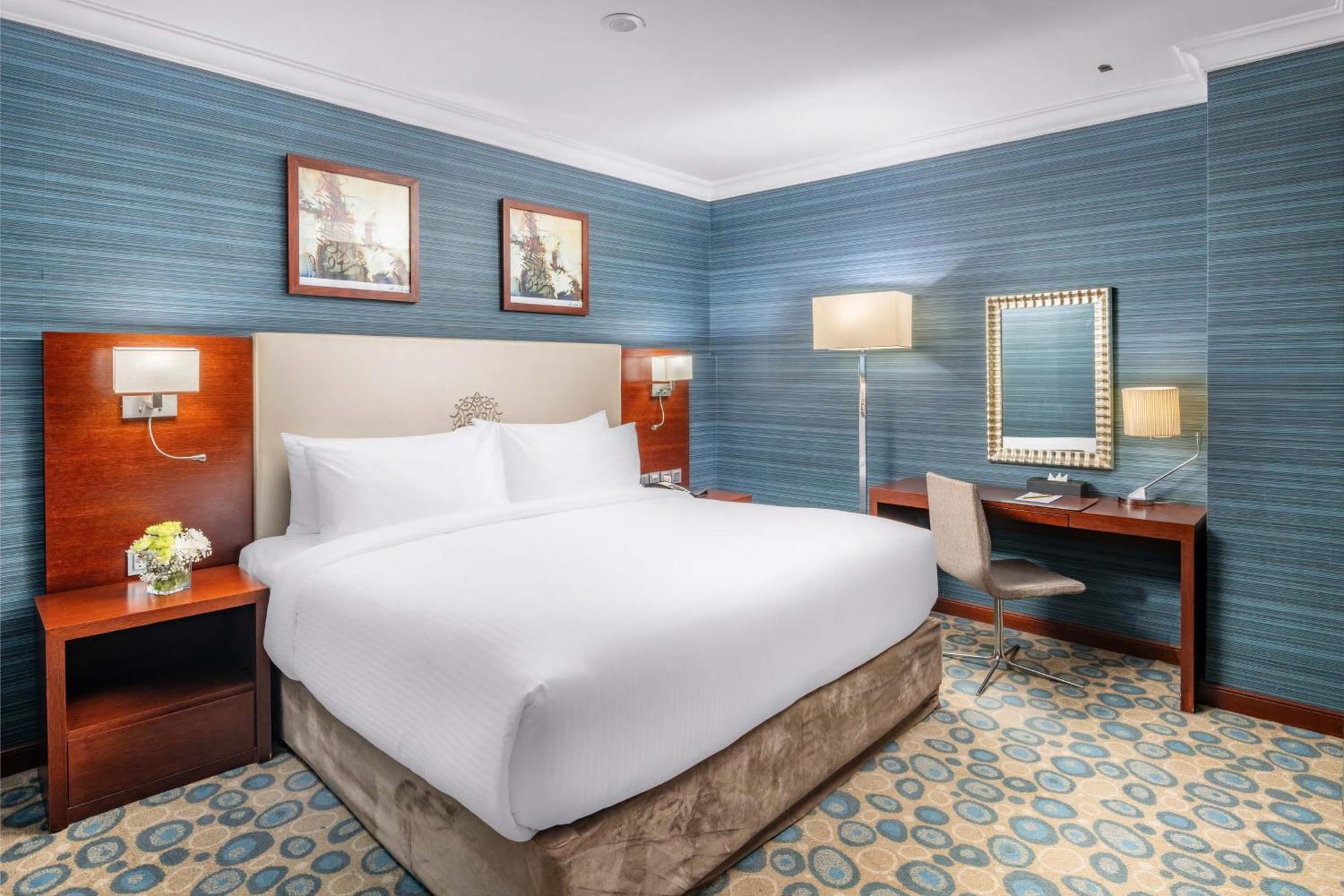 Al Aqeeq Madinah Hotel Medyna Zewnętrze zdjęcie The photo shows a well-furnished hotel room. It features a large bed with white linens, flanked by wooden nightstands. The walls are painted in a light blue color with a textured pattern. Above the bed, there are two framed pictures. On one side of t