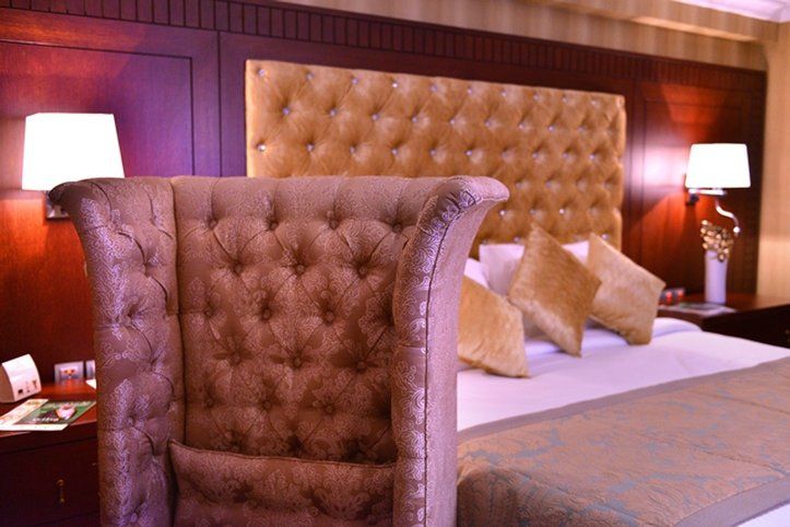 Al Aqeeq Madinah Hotel Medyna Zewnętrze zdjęcie The image shows a luxurious bedroom featuring a large upholstered headboard with a tufted design in a soft, beige color. The bed is elegantly dressed with decorative pillows and a textured bedspread. In the background, there are wooden paneling and s